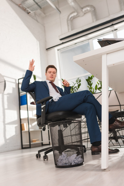 hombre de negocios guapo sentado en la silla y tirar papel en la papelera en la oficina, concepto de dilación
 - Foto, imagen