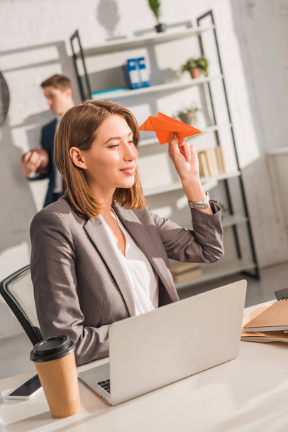 attractive businesswoman holding paper plane and sitting near laptop with coworker on background, procrastination concept - Foto, afbeelding