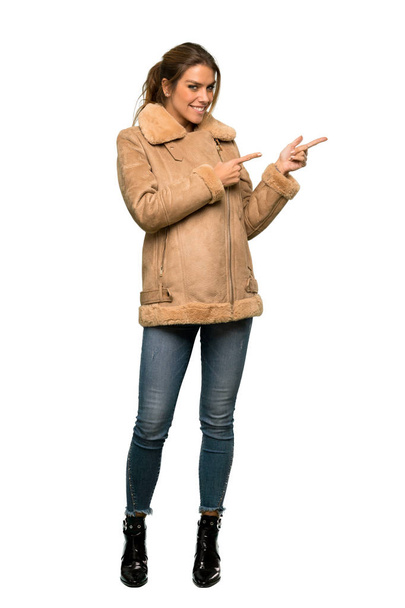 Una toma de cuerpo entero de una mujer rubia con un abrigo apuntando con el dedo hacia un lado sobre un fondo blanco aislado
 - Foto, Imagen