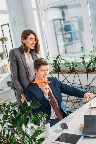 cheerful businesswoman standing near happy coworker holding paper plane in office  - Фото, изображение