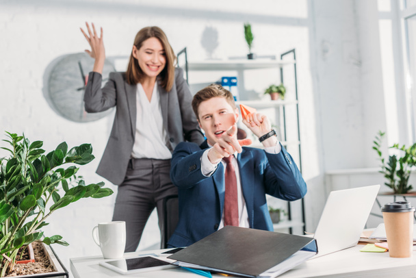 attractive businesswoman standing and gesturing near cheerful coworker holding paper plane and pointing with finger in office  - Фото, зображення