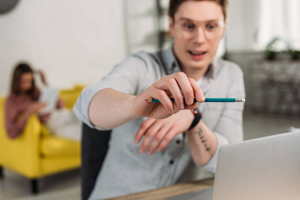 selective focus of man holding pencil near laptop with girlfriend on background - Фото, зображення