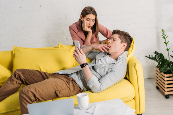 atractiva mujer mirando cansado novio usando teléfono inteligente y acostado en el sofá
 - Foto, imagen