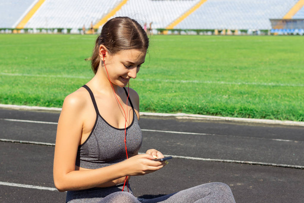 Красивая фитнес-девушка в серой спортивной одежде пользуется смартфоном и слушает музыку на стадионе после тренировки. Спорт и здоровая концепция
. - Фото, изображение