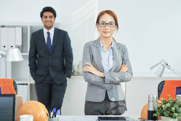 Portrait of young Asian female manager standing at office with arms crossed with businessman in background - Foto, afbeelding