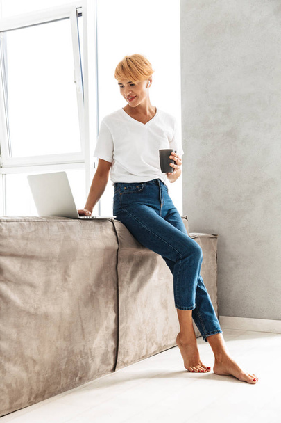 Pretty woman using laptop computer while sitting on a couch and drinking coffee - Foto, Bild