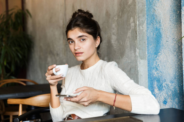 Photo of pretty serious young woman sitting in cafe drinking coffee indoors using mobile phone chatting. - Photo, Image