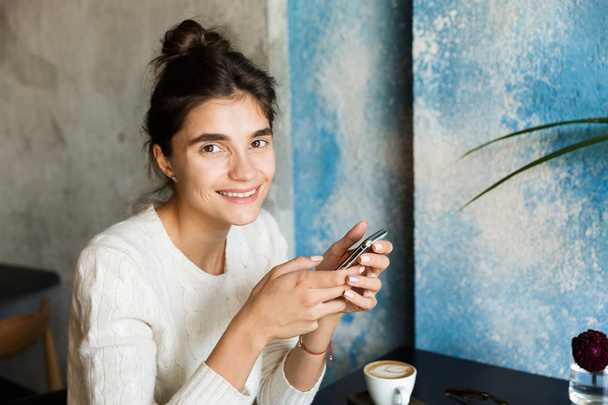 Image of pretty young woman sitting in cafe drinking coffee indoors using mobile phone chatting. - Photo, Image