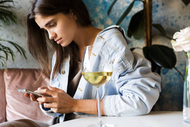 Photo of pretty beautiful young woman sitting in cafe indoors drinking white wine using mobile phone. - Фото, изображение
