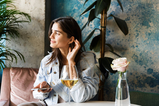 Photo of pretty beautiful young woman sitting in cafe indoors drinking white wine using mobile phone. - Foto, Imagen
