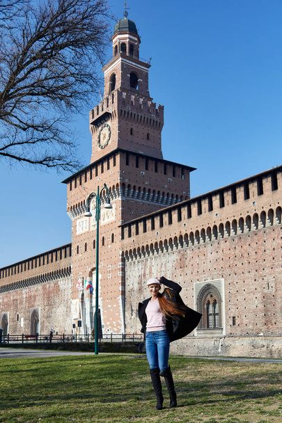 Милан, Италия - 18 февраля 2019 года Мода позирует модель с красным харом, шарфом и черным пальто для портрета с Castelo Sforzesco в Милане на заднем плане в солнечный день
 - Фото, изображение