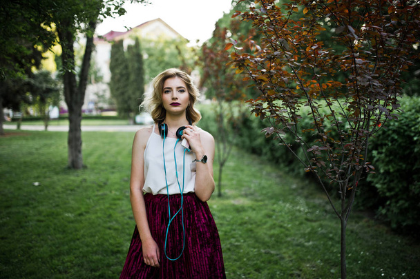 Fashionable and beautiful blonde model girl in stylish red velvet velour skirt, white blouse, posed at park with phone and earphones. - Foto, immagini