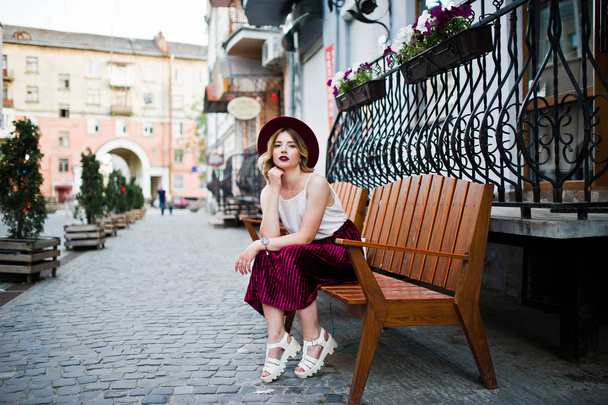 Fashionable and beautiful blonde model girl in stylish red velvet velour skirt, white blouse and hat, posed on bench at city street. - Valokuva, kuva
