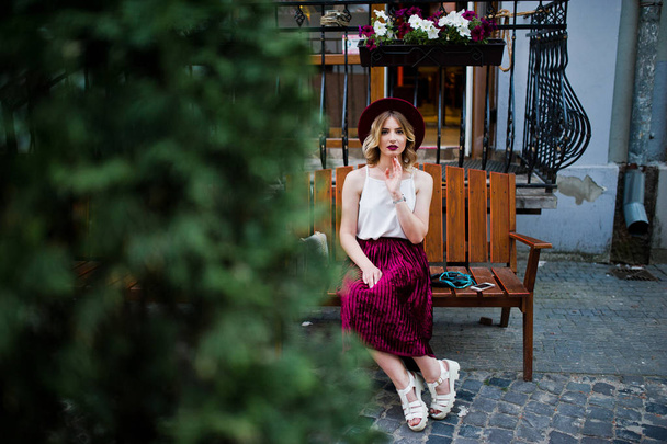 Fashionable and beautiful blonde model girl in stylish red velvet velour skirt, white blouse and hat, posed on bench at city street. - Photo, Image