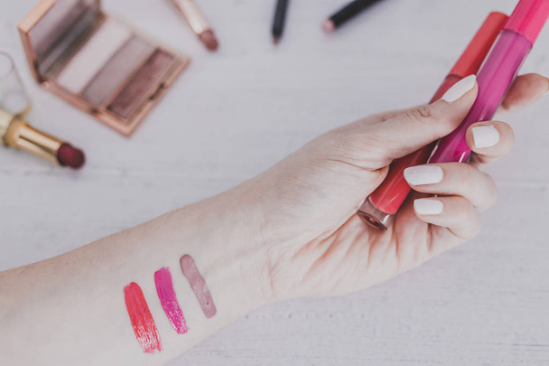 woman's hand holding lipgloss product and testing it surrounded by other make-up items on desk, concept of beauty industry - Foto, Imagen
