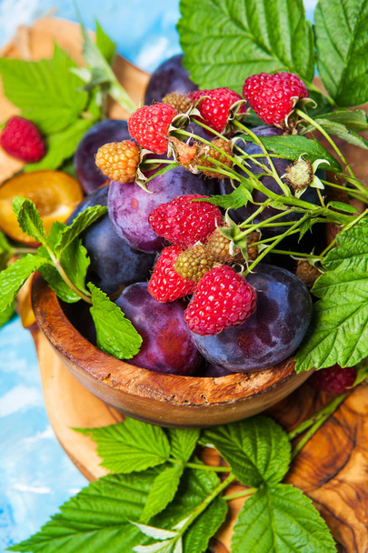 Closeup still life of plums and red raspberry with green leaves on wooden dish, summer autumn harvest  - Photo, Image
