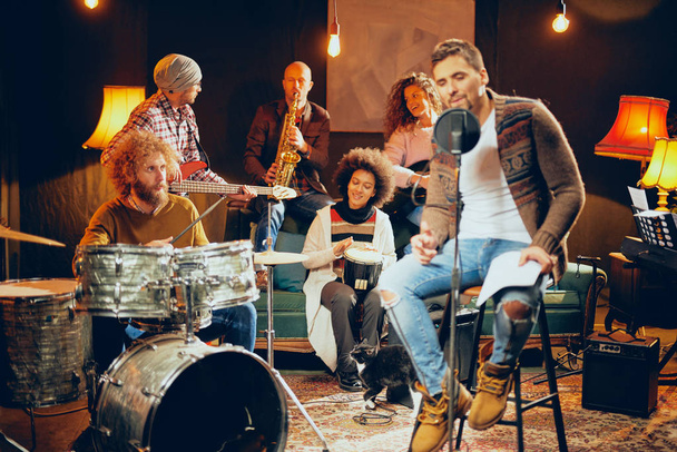 Homme chantant et assis sur une chaise tandis que son groupe jouait des instruments en arrière-plan. Accueil studio intérieur
. - Photo, image