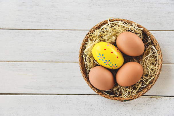 Easter egg and chicken eggs in basket nest decoration on white wooden background top view  - Photo, image