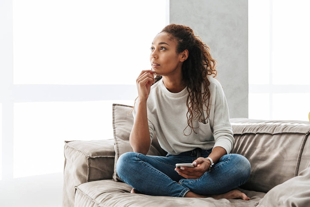 Photo of pretty african american woman using cell phone while sitting on couch in bright apartment - Photo, Image