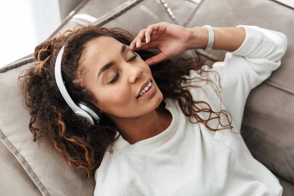 Photo of pleased african american woman listening to music using wireless headphones while lying on sofa at home - Zdjęcie, obraz