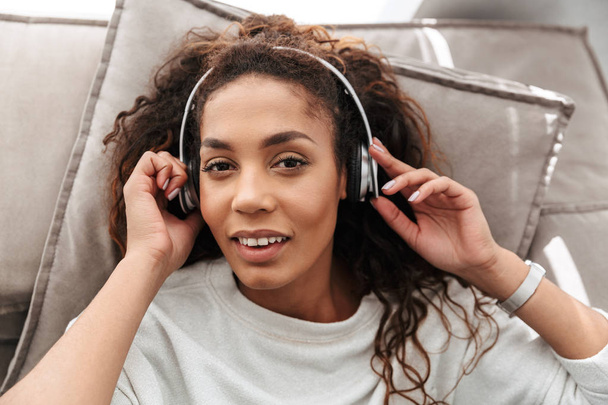 自宅のソファの上に横たわっている間無線のヘッドフォンを使用して音楽を聞いて魅力的なアフリカ系アメリカ人女性の写真 - 写真・画像