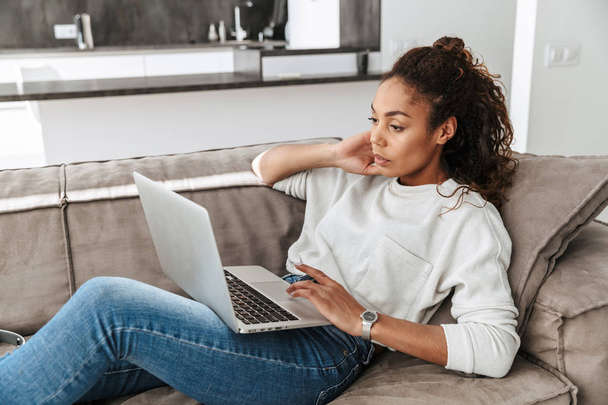 Photo of pleased african american woman using laptop while lying on sofa in bright living room - Φωτογραφία, εικόνα