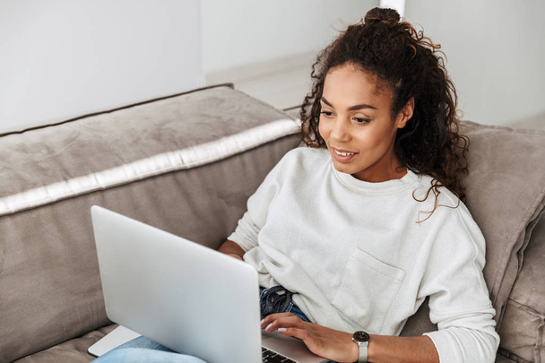 Photo of satisfied african american woman using laptop while lying on sofa in bright living room - Valokuva, kuva