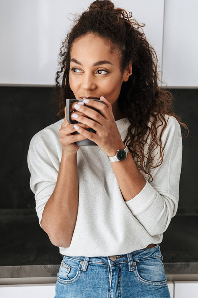 Image of happy african american girl drinking tea while standing in bright flat - Foto, Bild