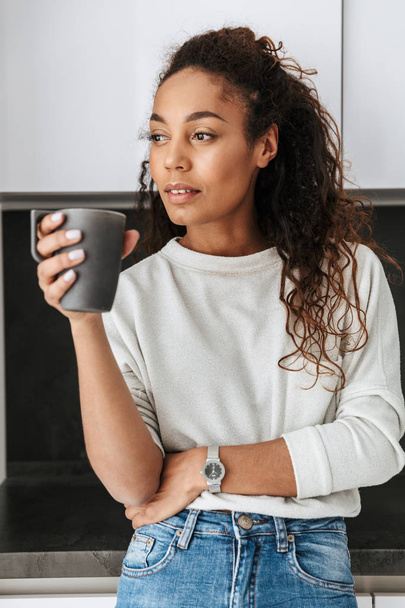 Image of adorable african american girl drinking tea while standing in bright flat - Valokuva, kuva