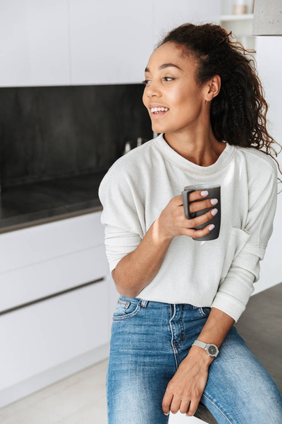 Image of smiling african american girl drinking tea while standing in bright flat - Foto, Bild