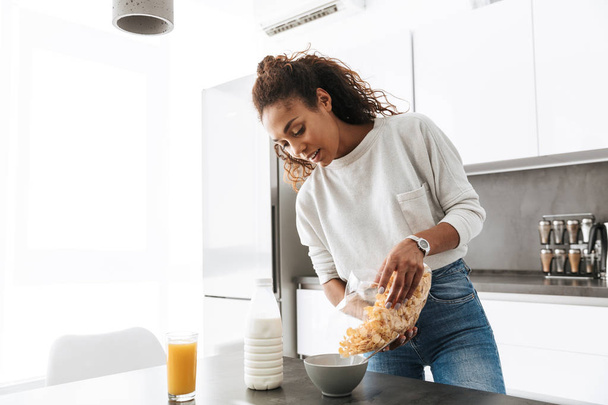 Image of smiling african american woman making breakfast with corn flakes and milk in stylish kitchen - Valokuva, kuva