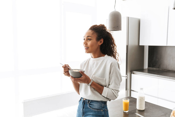Image of brunette african american woman eating corn flakes with milk on breakfast in stylish kitchen - Foto, afbeelding