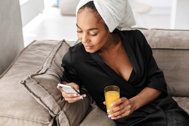 Portrait of pretty african american woman wearing housecoat using mobile phone and drinking juice at home - Фото, зображення