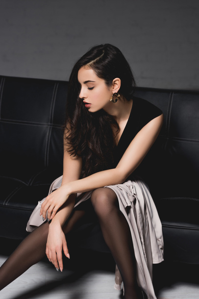 brunette woman in black dress sitting on couch on grey background - Zdjęcie, obraz