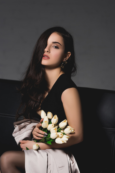 attractive brunette woman in black dress sitting on couch and holding white tulips on dark background - Fotó, kép