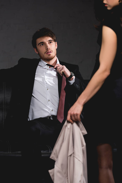 selective focus of handsome man sitting on couch and taking off tie wile brunette woman standing opposite on dark background - Photo, Image