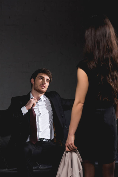 brunette woman standing opposite handsome man sitting on couch and taking off tie on dark background - Foto, afbeelding