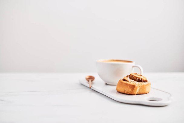Freshly baked cinnamon roll with spices and cocoa filling and coffee or cappuccino on white serving plate on white marble table over white background. Swedish breakfast. Copy space. Selective focus. - Φωτογραφία, εικόνα
