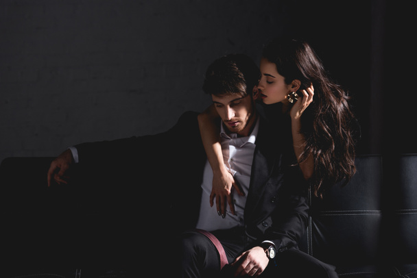 brunette woman in black dress standing behind couch, hugging and kissing handsome man in suit sitting on black background  - Foto, Imagen