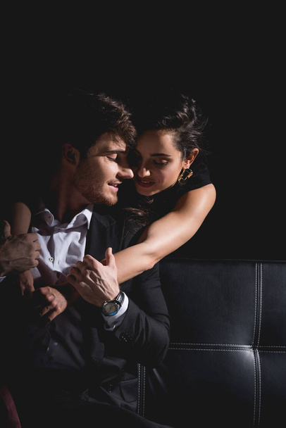attractive woman standing and hugging handsome man sitting on couch isolated on black - Photo, Image