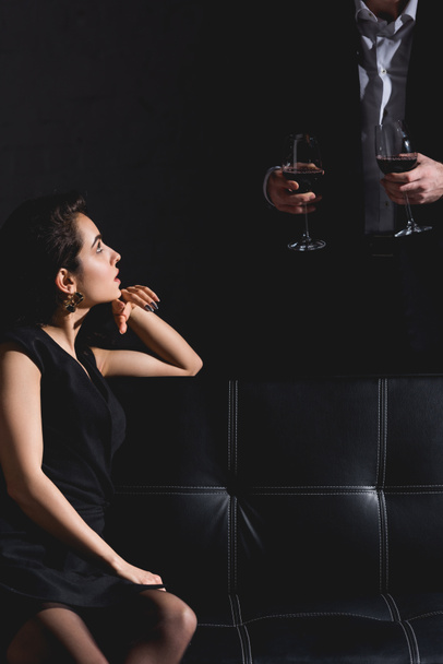 brunette woman sitting wile handsome man in black suit standing behind couch and holding glasses with wine on black background - Photo, Image