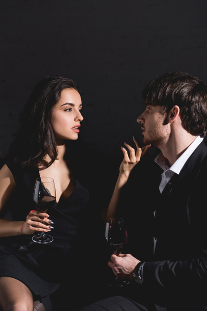 attractive woman in dress and handsome man in black suit sitting on couch with glass on black background - Photo, Image