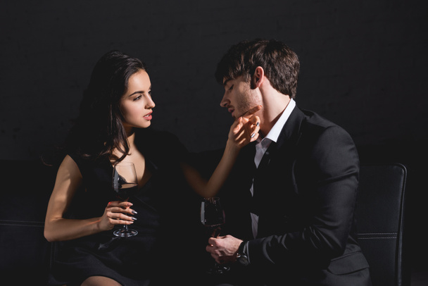 attractive brunette woman in black dress and handsome man in suit sitting on couch with glass on black background - Foto, Bild