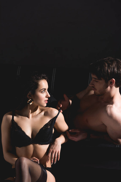 handsome man lying on couch and undressing woman in lingerie sitting on floor on black background  - Фото, изображение