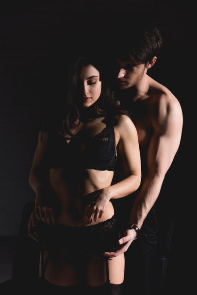 attractive brunette woman in black lingerie and shirtless handsome man standing and hugging isolated on black - Фото, изображение
