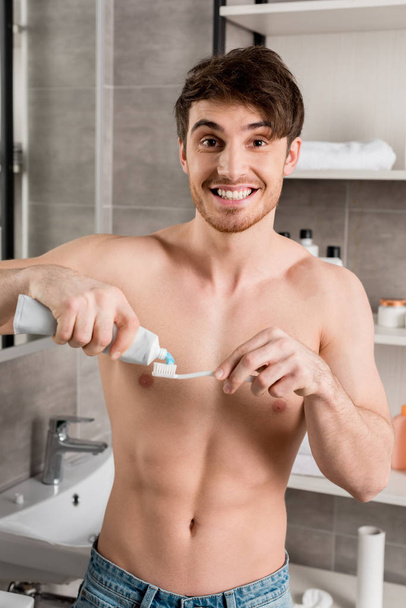 handsome shirtless man putting toothpaste to toothbrush in bathroom - Foto, Imagen