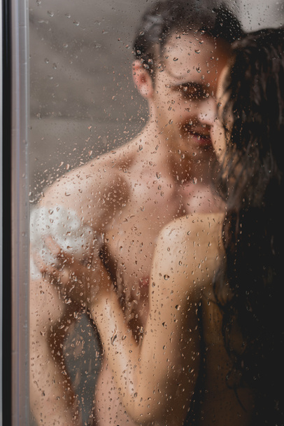 Selektiver Fokus des sexy Paares beim Umarmen und Duschen mit Luffa in der Kabine  - Foto, Bild