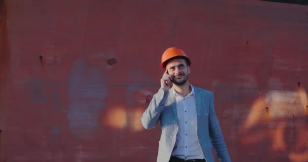 engineer in grey business suit and orange helmet speaks on phone looks at wrist watch against brown blurry wall - Πλάνα, βίντεο