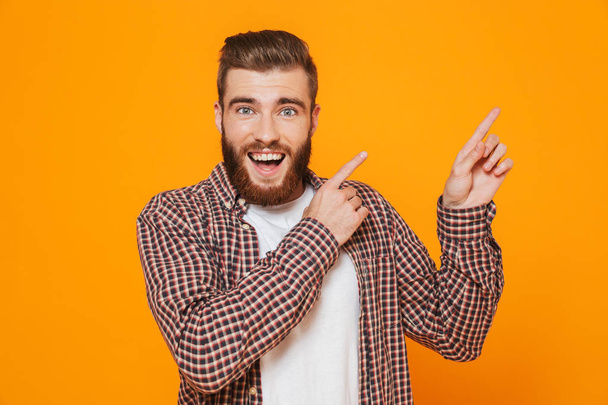Portrait of a cheerful young man wearing casual clothes standing isolated over yellow background, pointing away at copy space - Foto, afbeelding