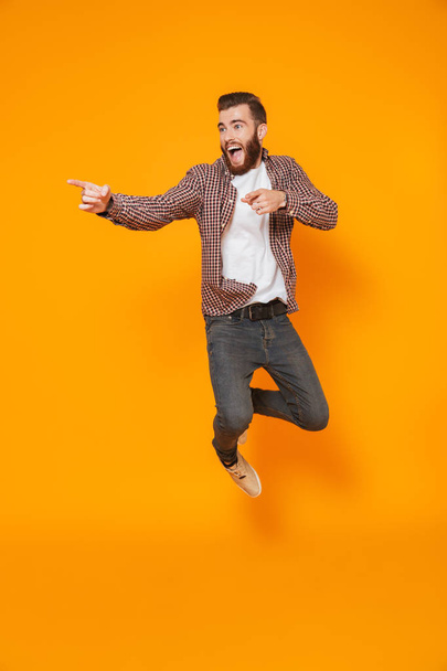 Full length portrait of a cheerful young man wearing casual clothes isolated over yellow background, pointing away at copy space, jumping - Valokuva, kuva
